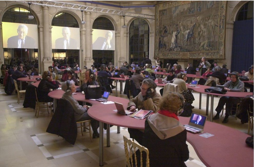  Salle avec des tables et des personnes assises aux tables, écoutant une conférence sur de petits écrans.