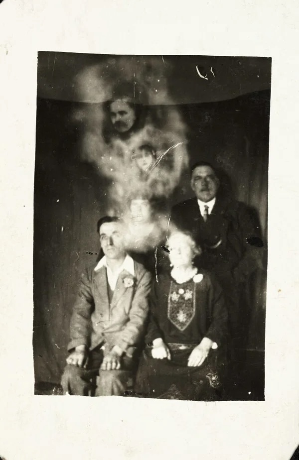 Photographie en noir et blanc. Quatre personnes posent, à la manière d'une photo de famille. En surimpression, plusieurs visages apparaissent au-dessus des quatre corps assis, comme s'il s'agissait de spectres.
